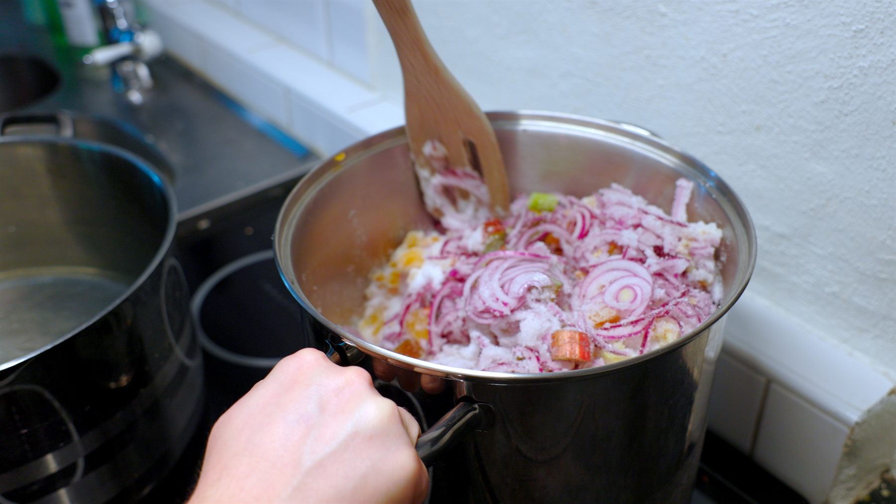 Rabarber, rödlök och socker ligger i en kastrull och rörs om med trägaffel.